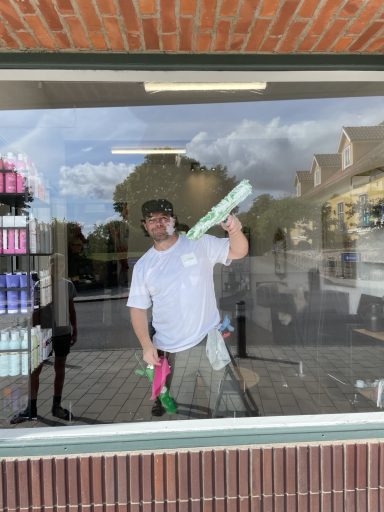 Man cleaning a shop window with spray bottles, standing outside in daylight.